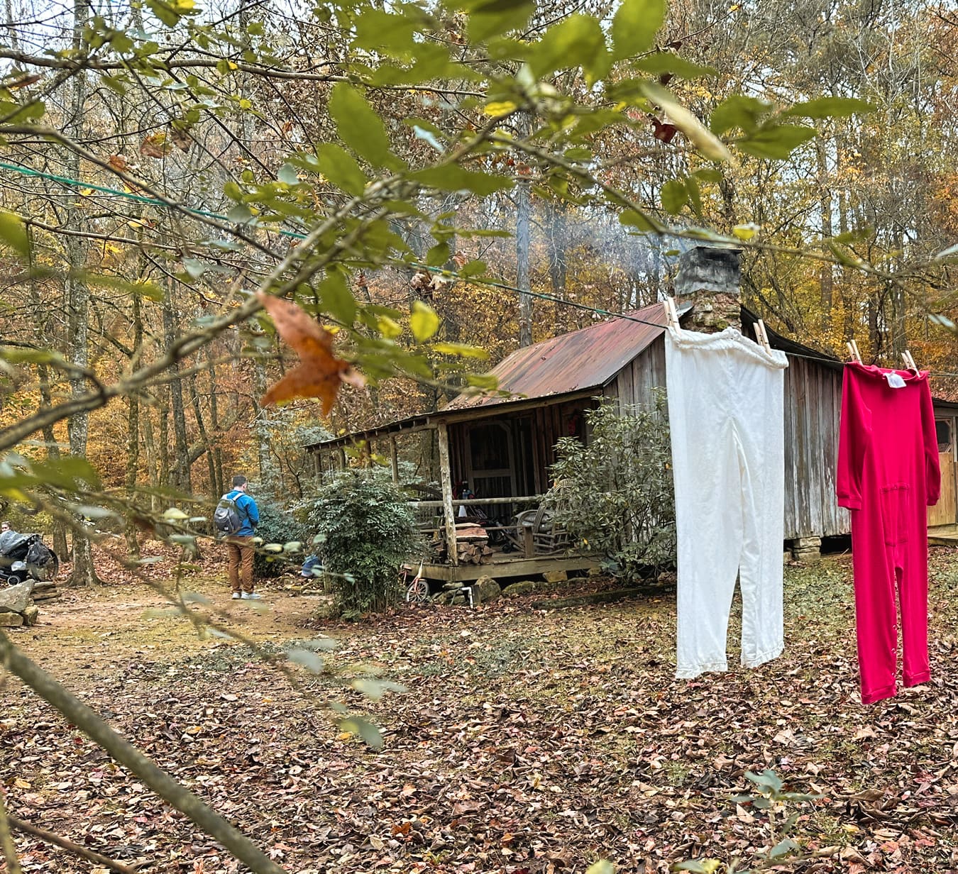A man standing in front of a cabin from the 1800s with smoke coming out of the chimney. In the foreground are two items of clothing hanging on a line. Fall colors on the leaves.