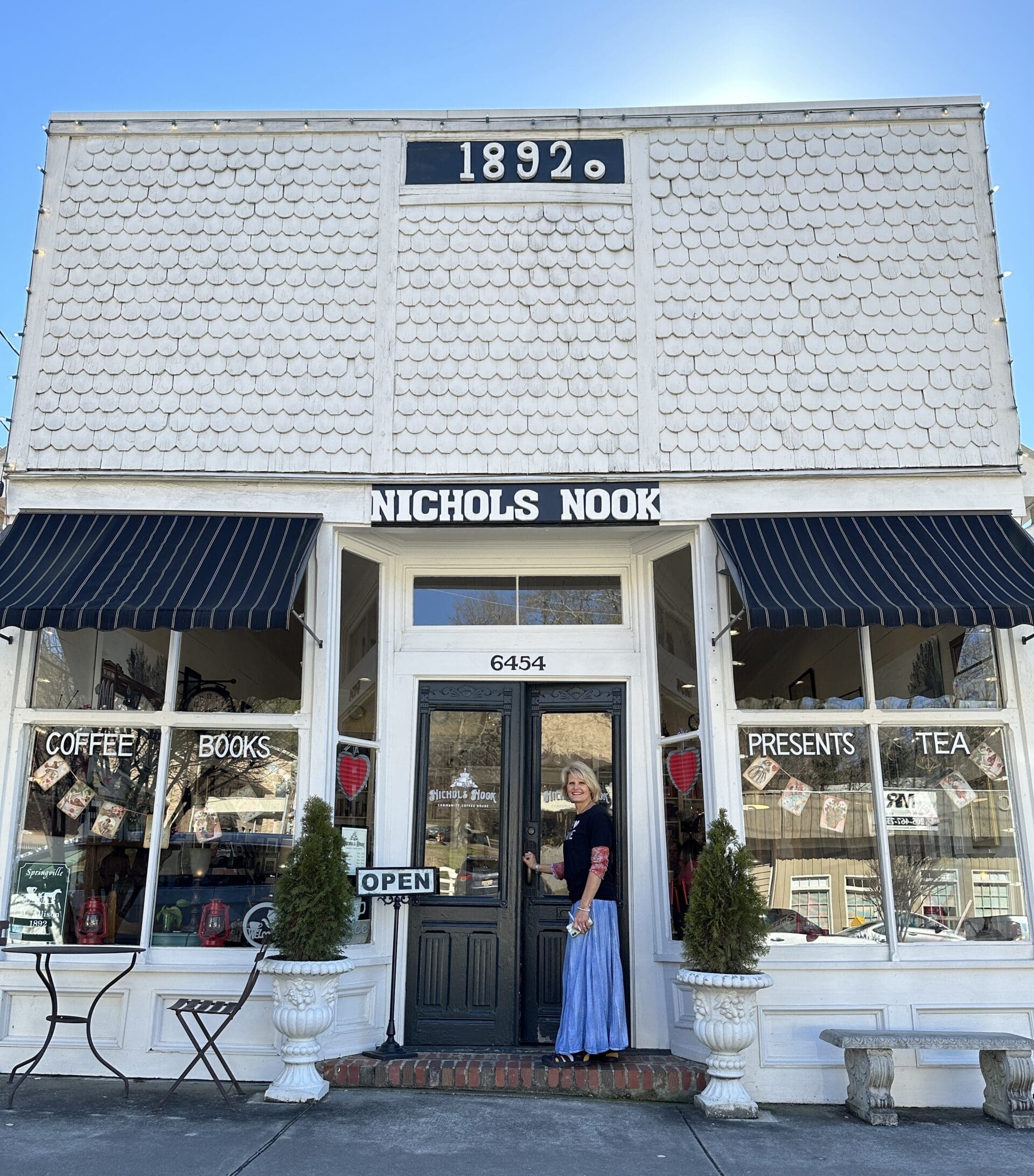 Woman standing in front of historic building with big windows. Sign at the very top reads "1982" and the sign above the door reads "Nichols Nook".