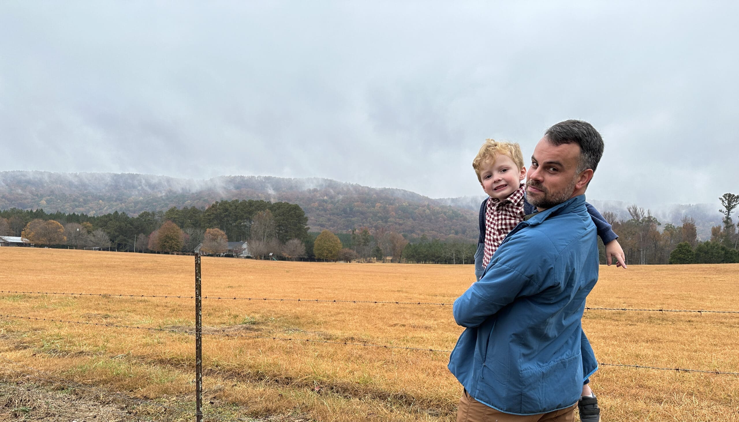 Man holding a young child in front of a fence, mist in the distance on Pine Mountain in Alabama. Late Fall colors.