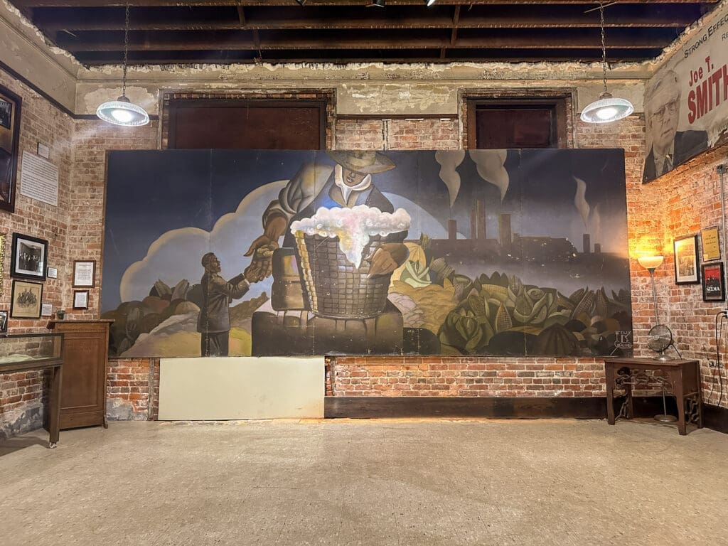 Large mural housed inside large brick building. Mural features a Black man in a suit and a Black farmer exchanging peanuts. Background: a farm scape on the left and a factory scape in the background on the right. Presumably the mural is a reference to George Washington Carver.
