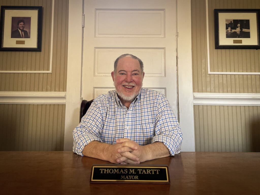 A man sits at a desk smiling with a nameplace in front of him reading "Mayor Tom Tartt".
