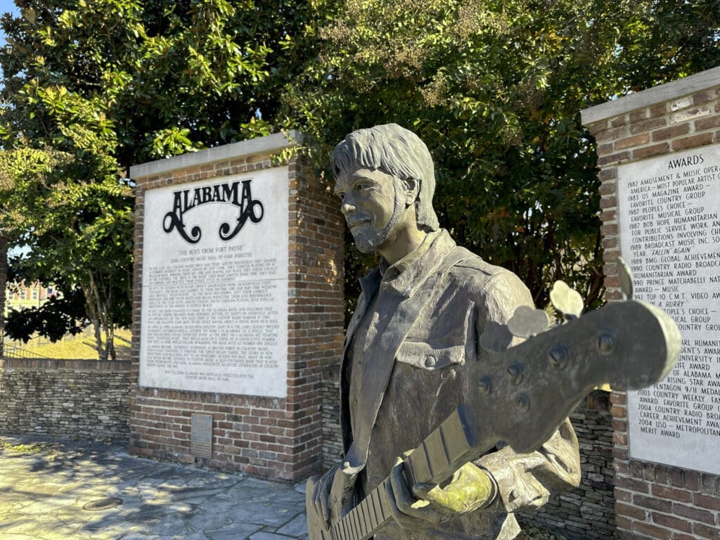 Foreground: a statue of a man playing guitar. Background: signage featuring the band Alabama's logo at top with paragraphs of text underneath.