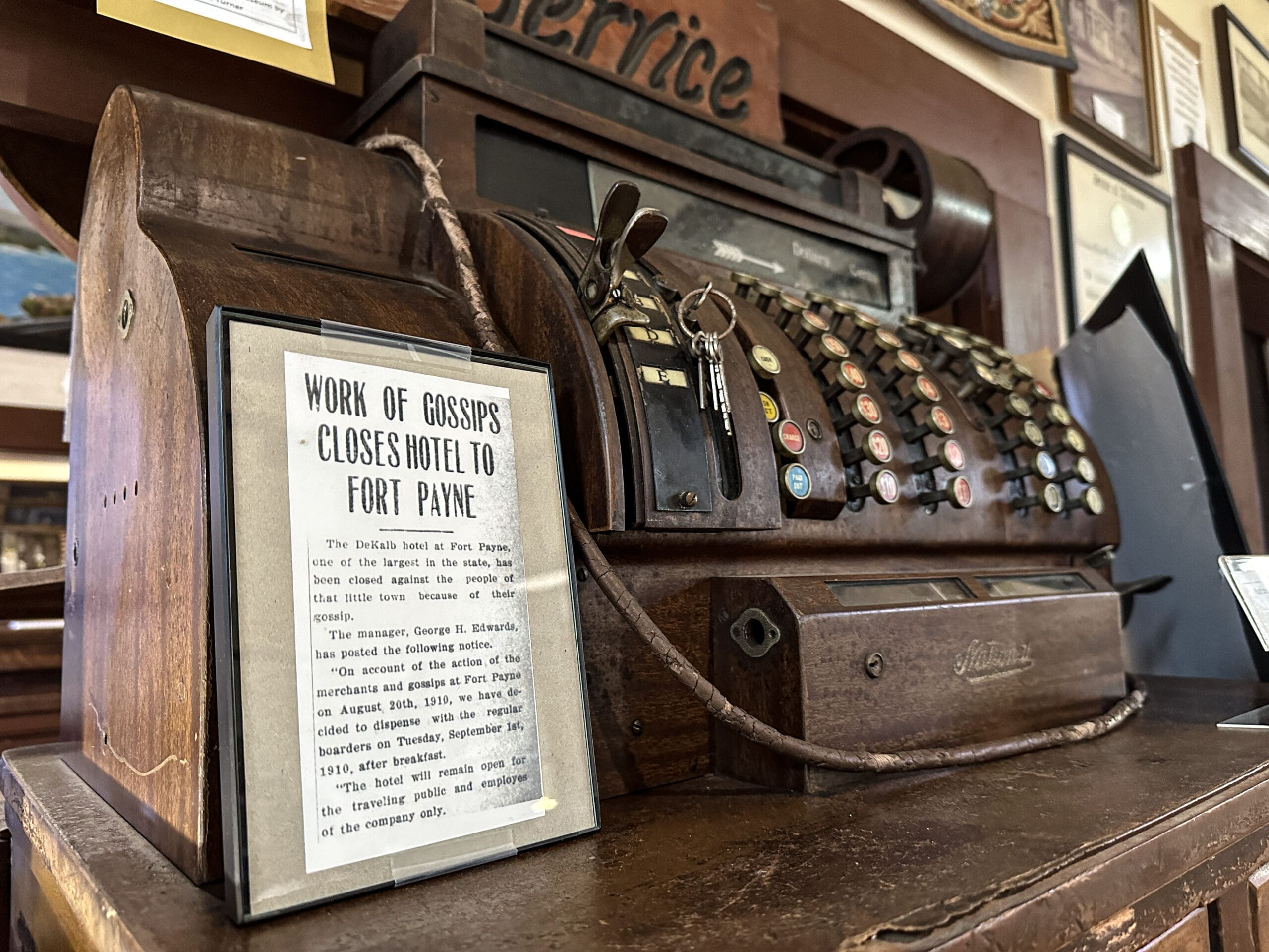 A vintage cash register with a framed newspaper clipping in front. Newspaper reads: "Work of Gossips Closes Hotel in Fort Payne".