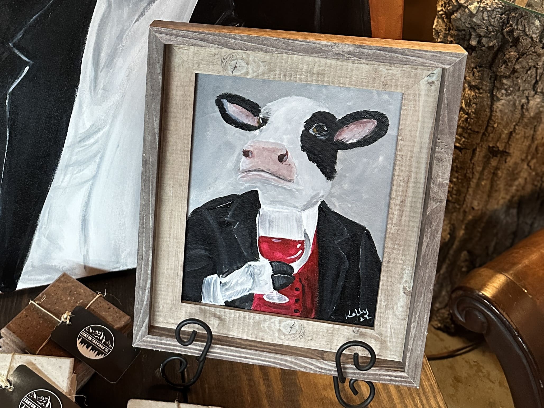 A wood-framed painting of a cow holding a glass of red wine.