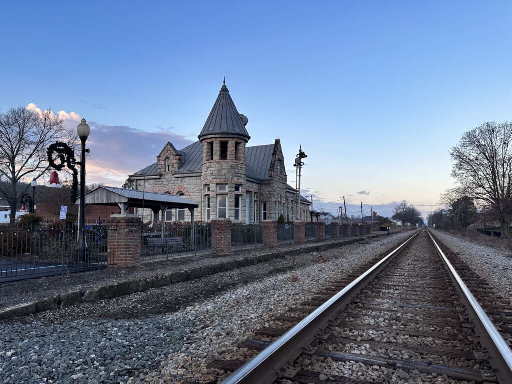 A historic stone railroad depot with a spire next to a railroad. Photo taken at sunset.