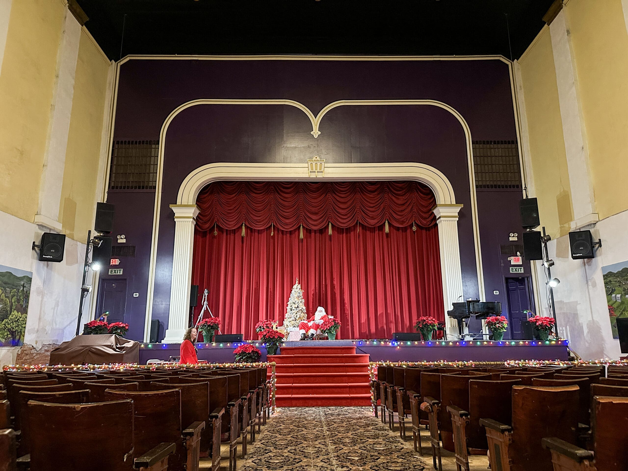 Interior of historic opera house featuring seating, a stage with a red curtain. A man dressed as Santa is sitting on the stage next to a Christmas tree.