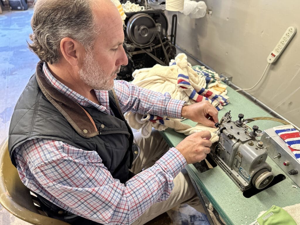 A man siting and sewing together the end of a pair of socks.