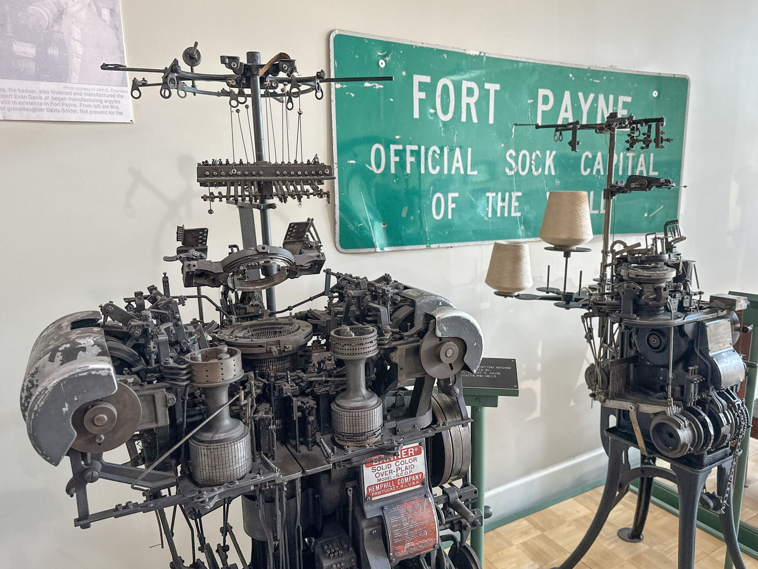 Complex vintage sock knitting machines with a large sign on the wall in the background reading "Fort Payne: Official Sock Capital of the World".