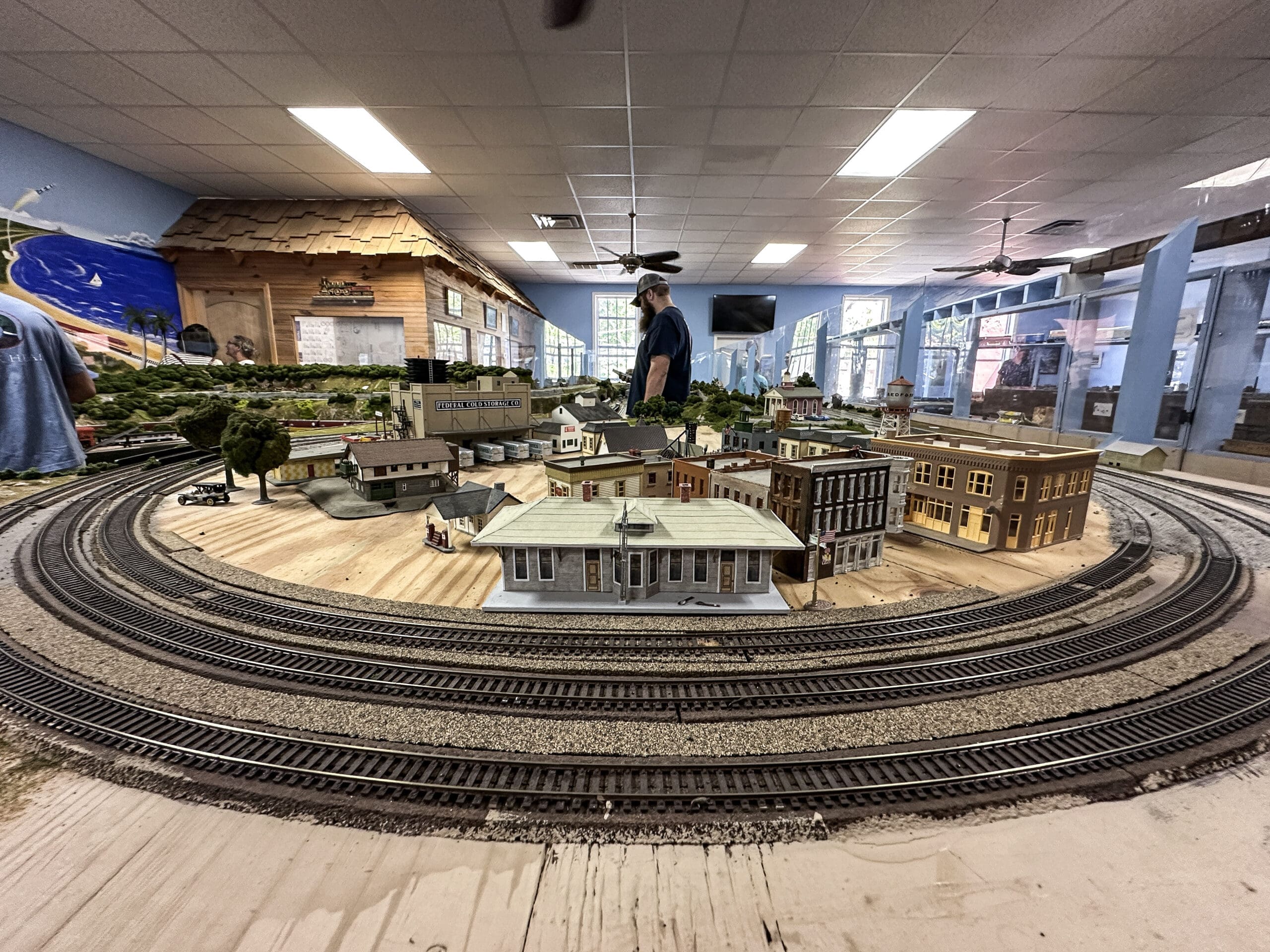 A large model train track in the foreground with a recreated model historic downtown. In the background, a person works on the trains.