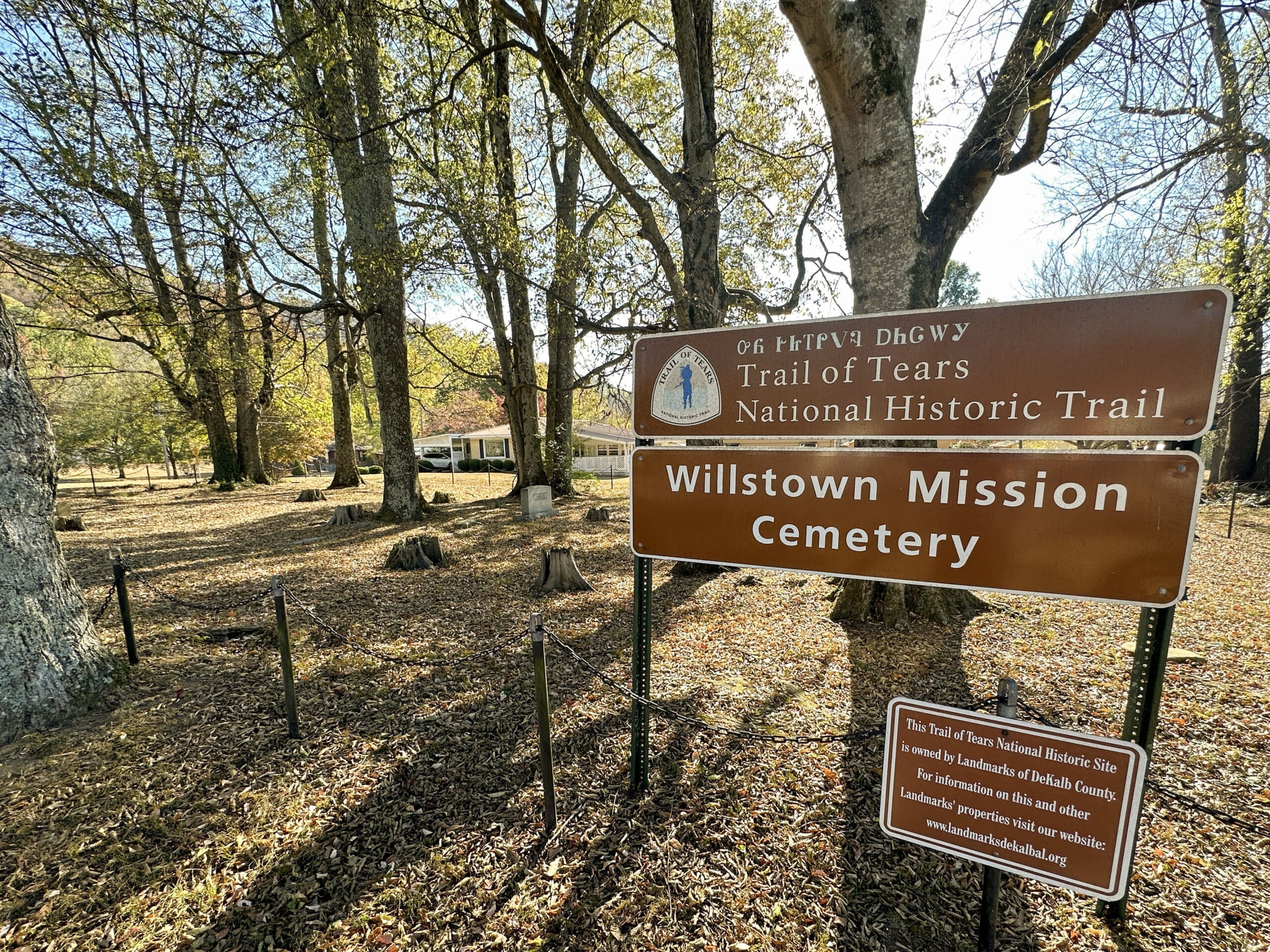 A sign in front of an old, small cemetery reads "Willstown Mission Cemetery" with writing in Cherokee as well.