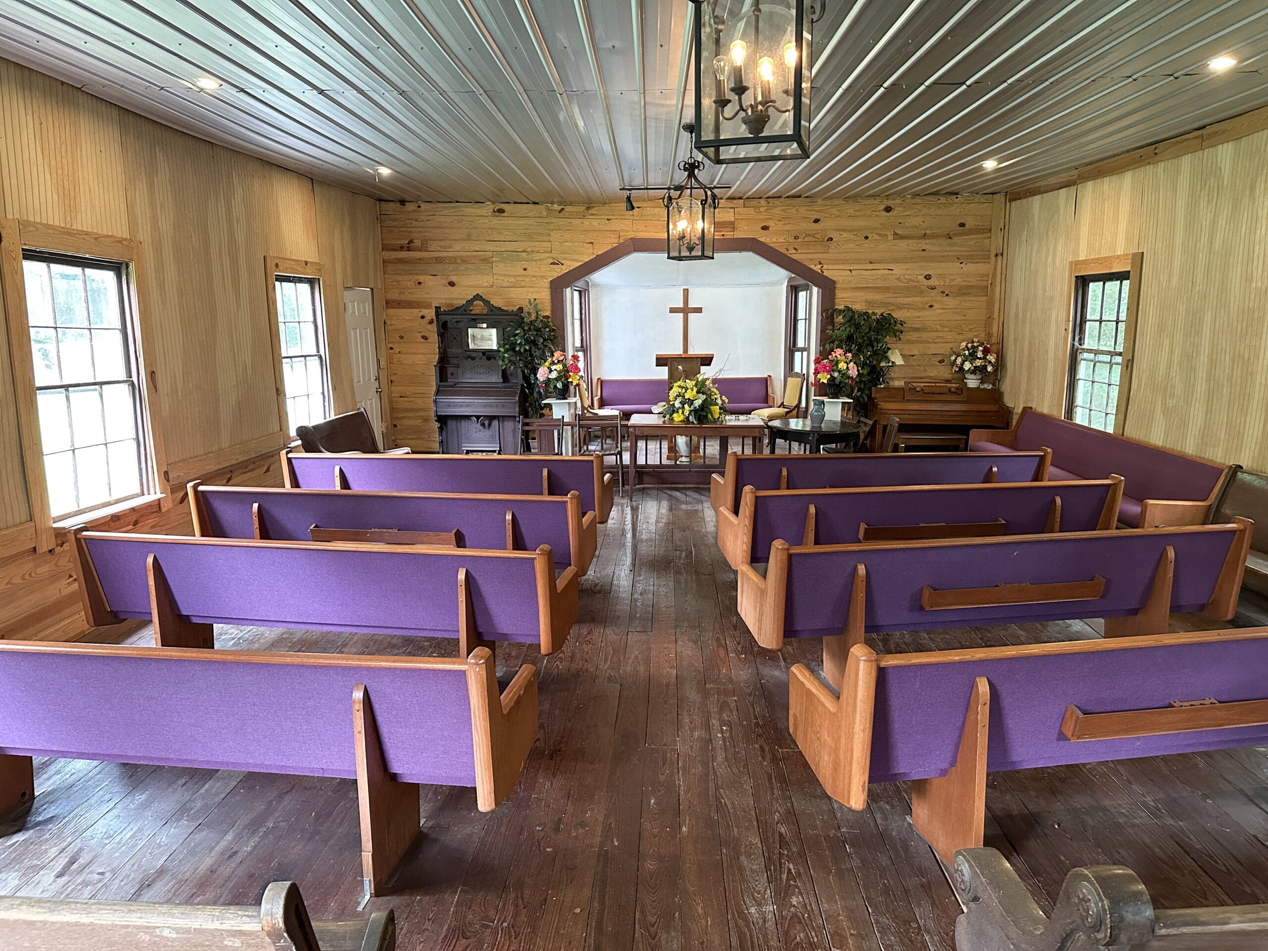 Inside view from the rear of a small, historic rural church. Two rows of pews with purple upholstery lead up to an altar.