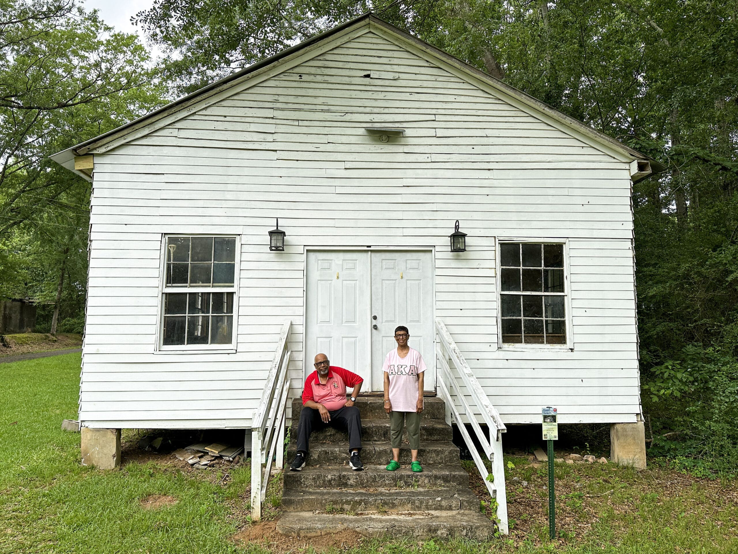 A black couple in front of a small, rural historical black church with white siding.
