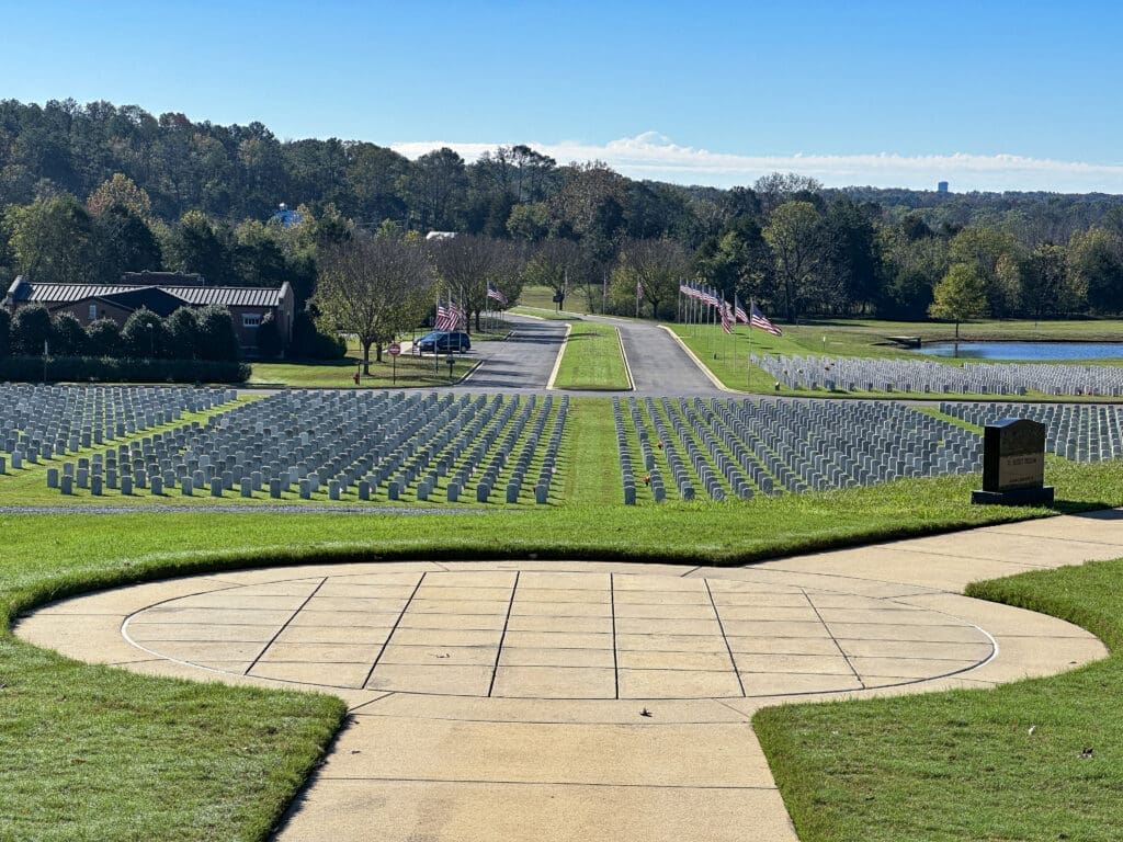 A concrete viewing point overlooking graves at a military cemetery. In the far background there is a road lined with flags and a small lake visible to the right.