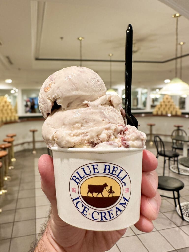 A hand holding a cup with multiple scoops of blackberry cobbler ice cream. Cup features the Blue Bell Ice Cream logo.