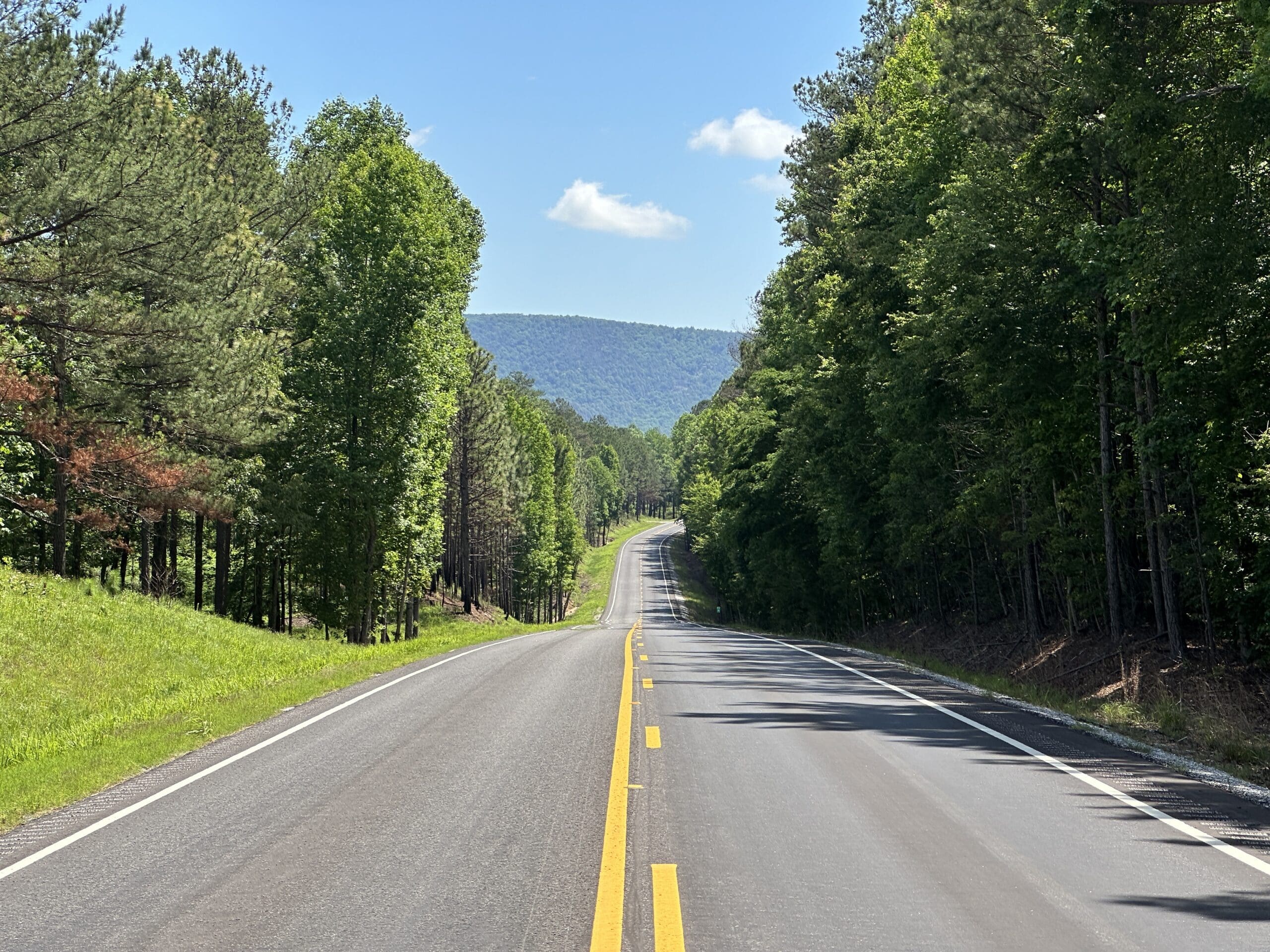 A highway with no cars and trees on each side heads off into the distance. Above the treeline, a small Appalachian mountain is visible.