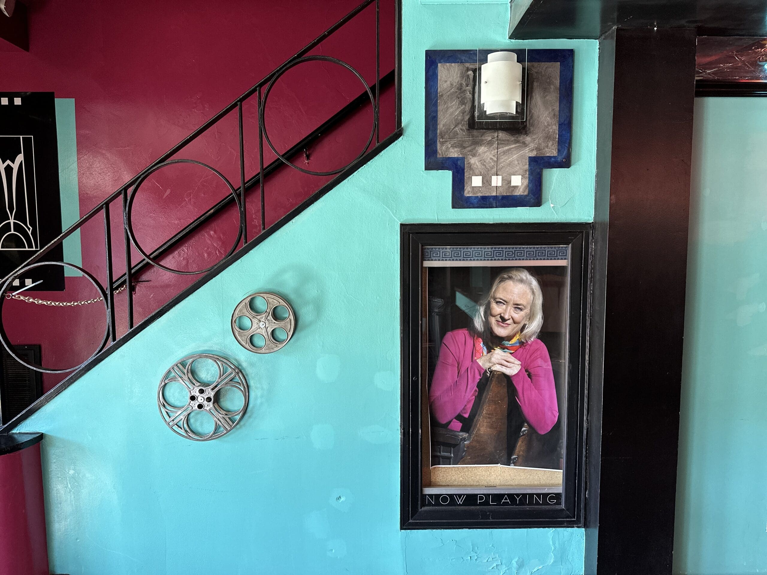 A portrait of an older actress hangs beside a staircase in a historic theatre with walls painted light blue and crimson.