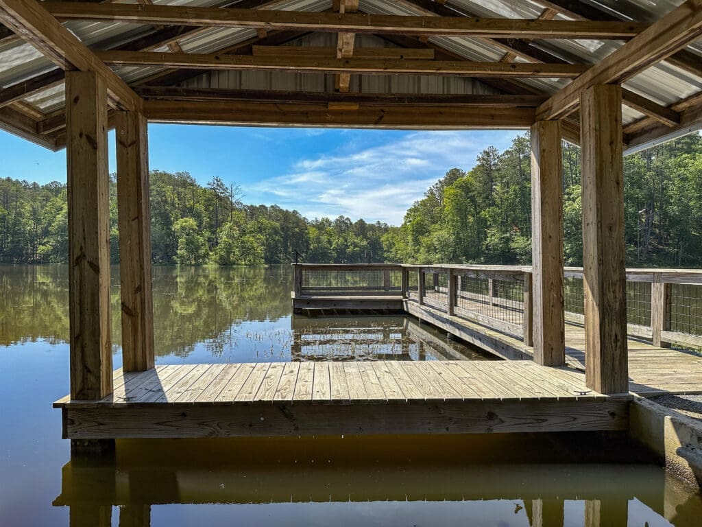 View from under roof of a covered dock. A pier runs along the right side ending in the middle ground of the photo. Background: green trees around a lake.