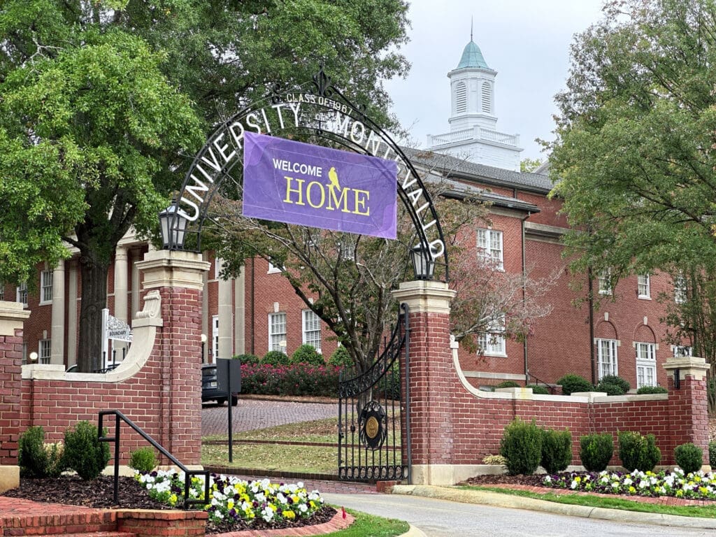 A brick-walled, flower-decorated entrance to a university. An archway over the entrance reads "University of Montevallo" while a hanging sign reads "Welcome Home". Green-leafed trees are visible and a third-story cupola sits high in the background.