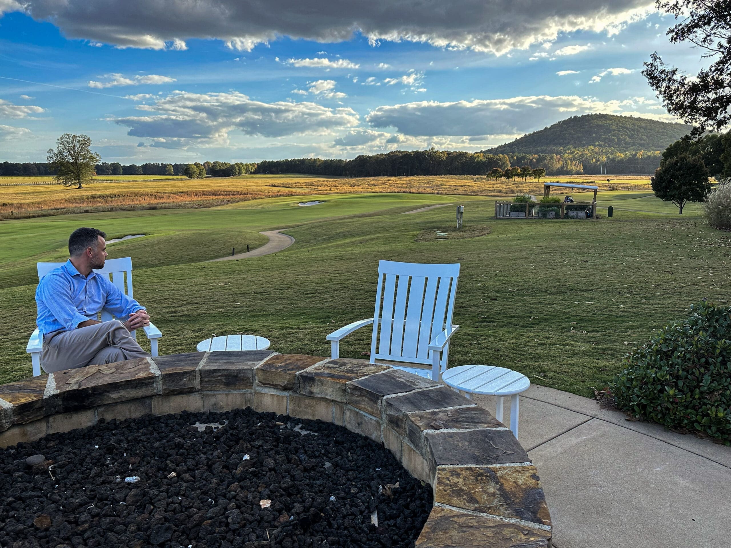 A man sits in front of a firepit looking off towards the sun setting over a low hill.