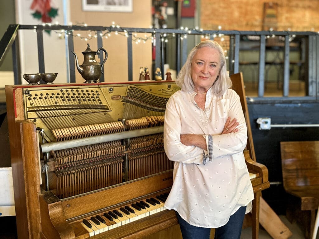 An older woman stands with her arms crossed, leaning against a vintage piano.