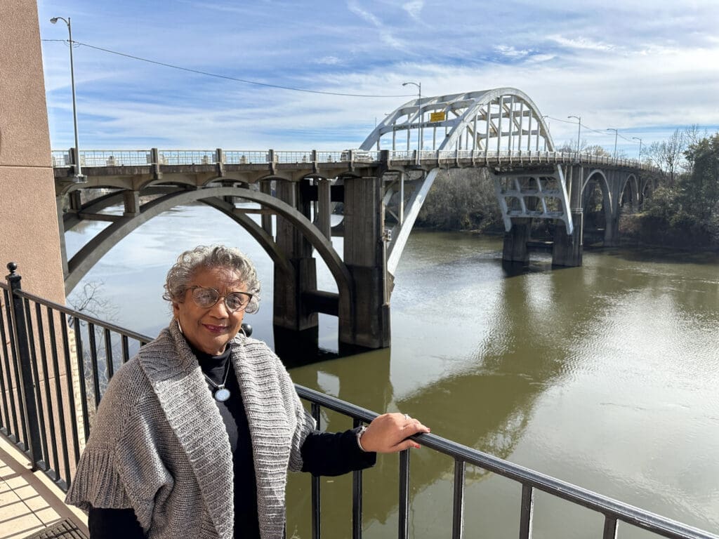 An older Black woman with her hand on a railing over a river. In the background, Selma, AL's famous Edmund Pettus Bridge.