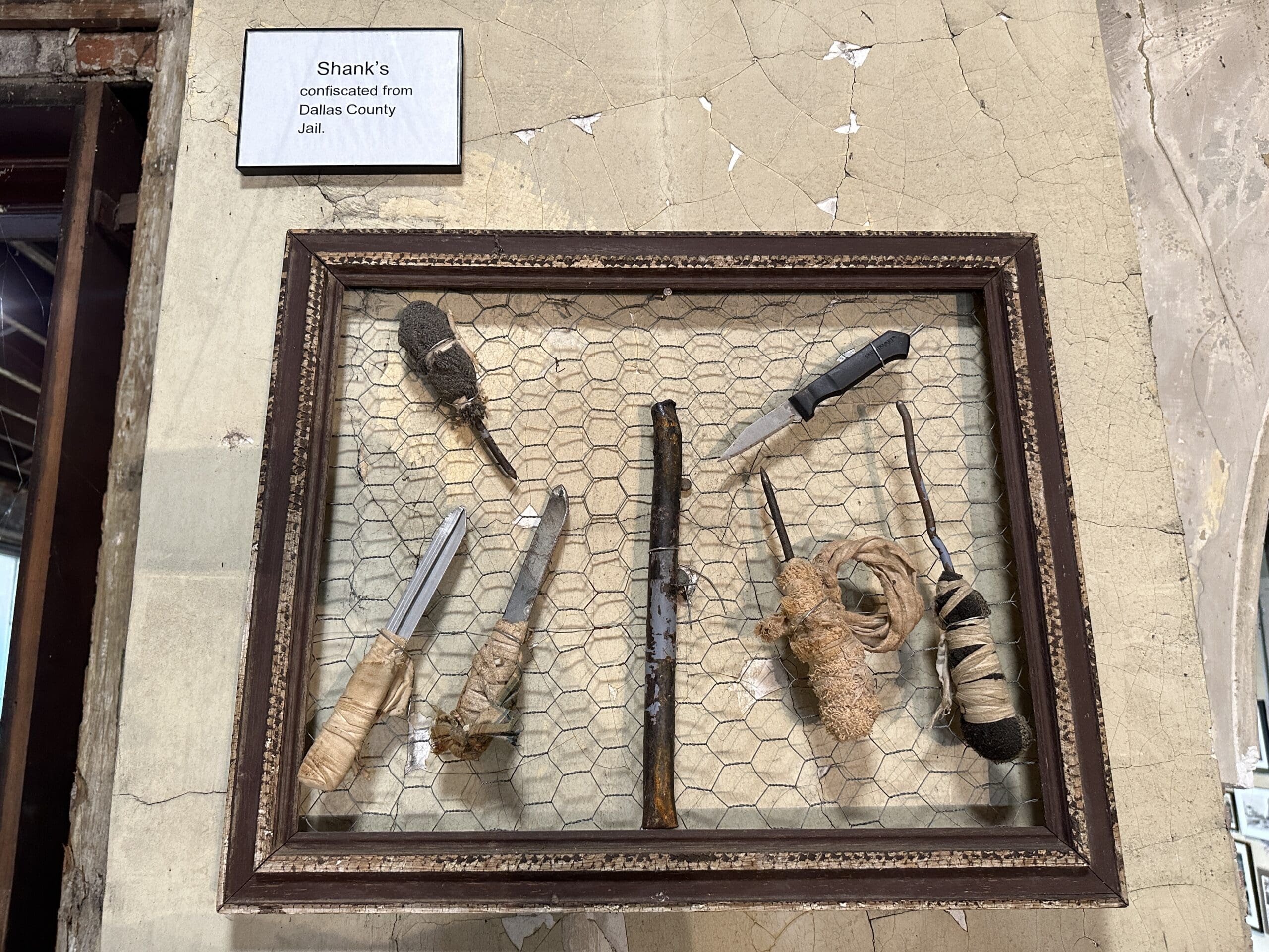 Multiple handcrafted prison shanks on chicken wire frame and hung on a wall. The sign above reads "Shanks confiscated from Dallas County Jail".
