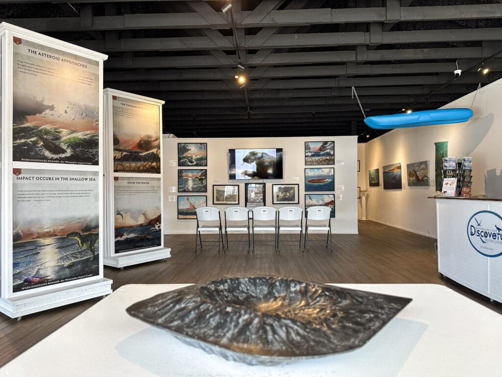 Interior of a visitors center with various informational signs, a TV, a kayak hanging from the ceiling and, in the foreground, a model of a meteor crater.